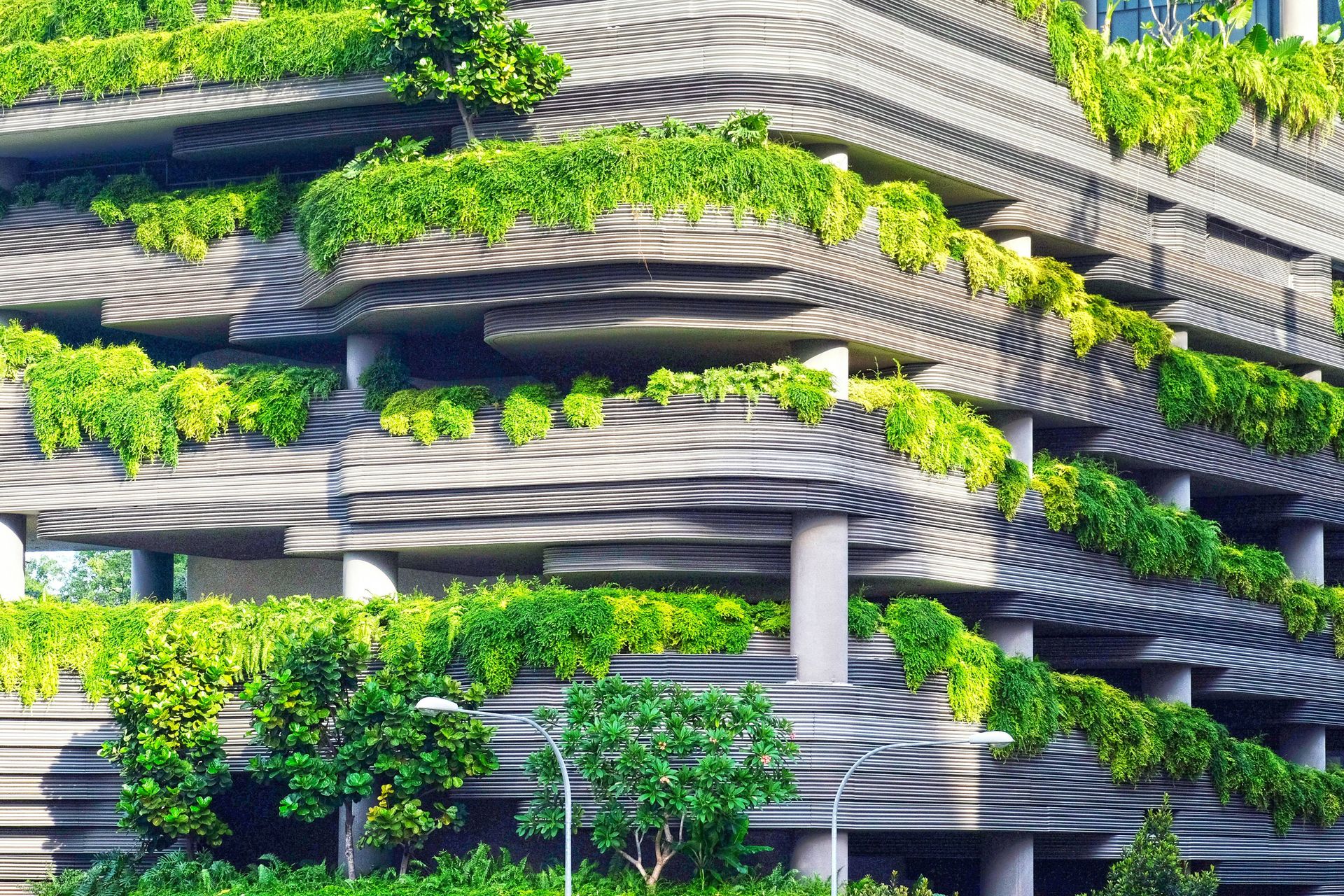 A close-up of a modern multi-story building featuring undulating concrete balconies overflowing with lush green tropical plants and trees.