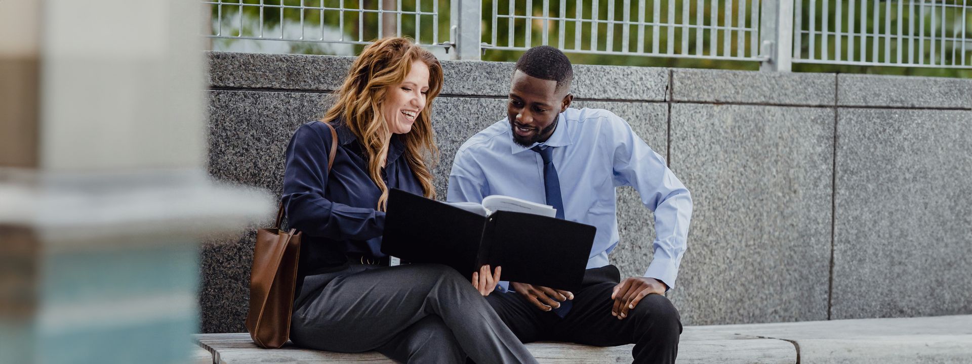 Two colleagues in professional attire sitting on an outdoor bench and smiling while reviewing a document together.