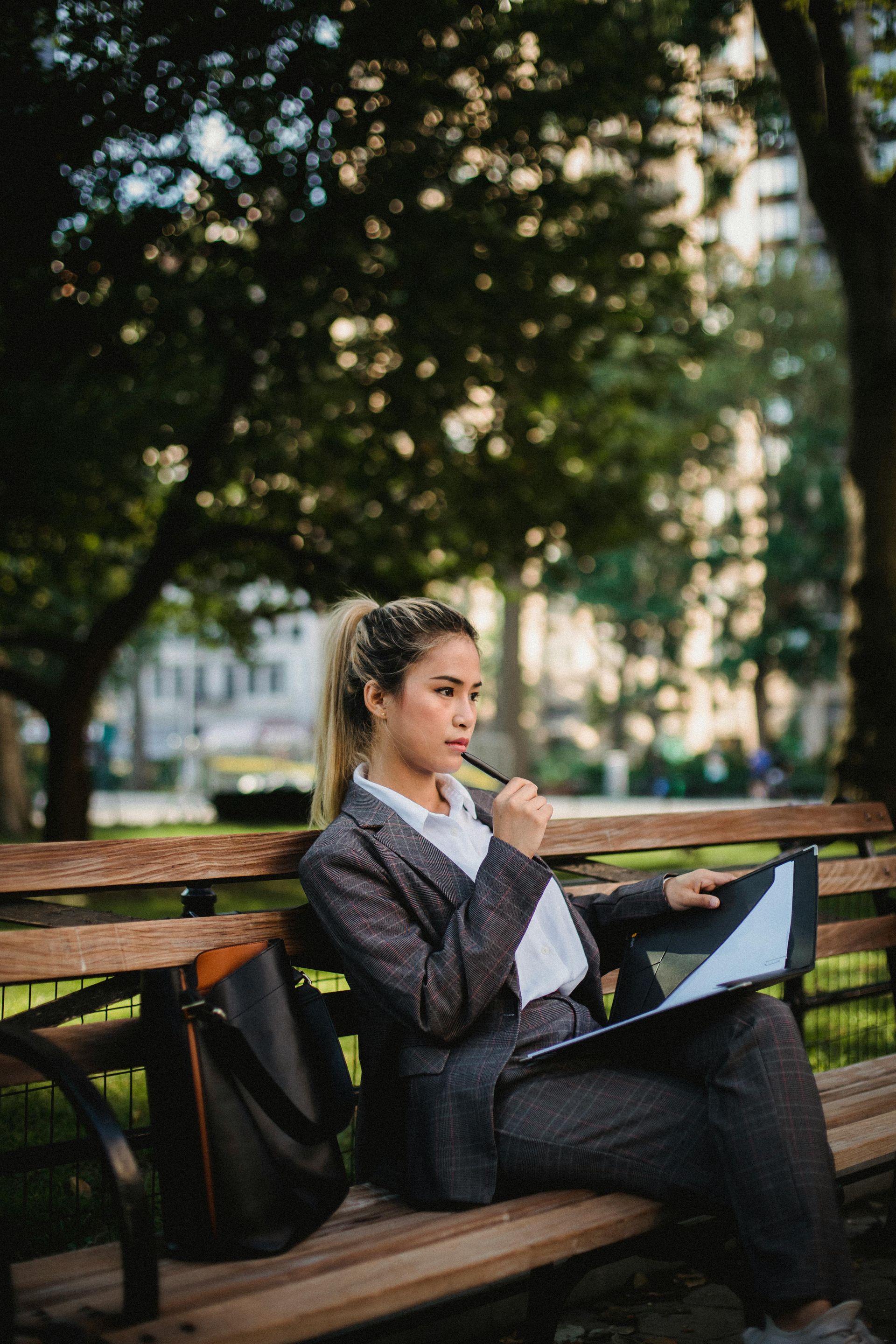 Focused woman sitting outdoors with a folder and pen, surrounded by green trees.